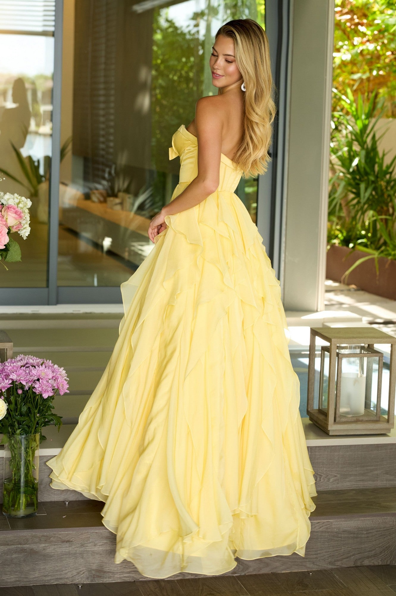 A woman wearing the Ava Presley 42209 Strapless Ruffled Prom Gown stands on outdoor steps, her floor-length yellow dress surrounded by flowers and lanterns in a magical evening scene.